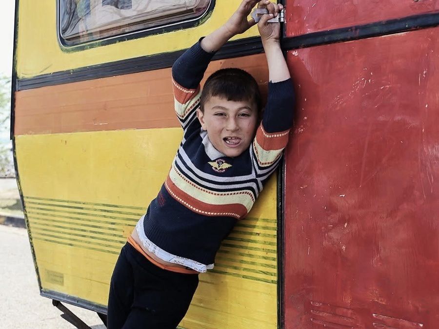 Portrait of a Boy, Idomeni, Greece: a boy hanging from the door of a red and yellow caravan, pulling a face. Photograph by Marcus Vallance.
