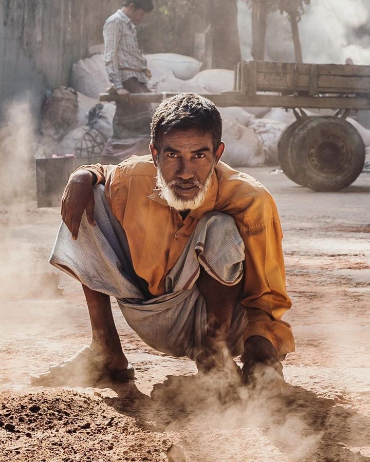 Street Worker, Bangladesh: a man in an orange shirt crouching in drifting dust. Photograph by Marcus Vallance.