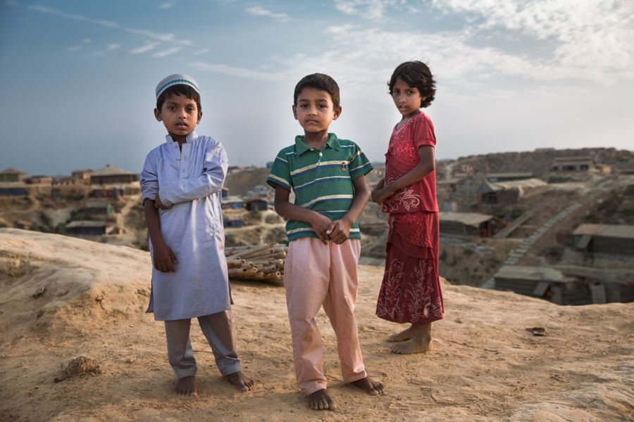 Three Siblings, Kutupalong Refugee Camp, Bangladesh: three children standing barefoot on a sandy ridge above the shelters of the camp. Photograph by Marcus Vallance.