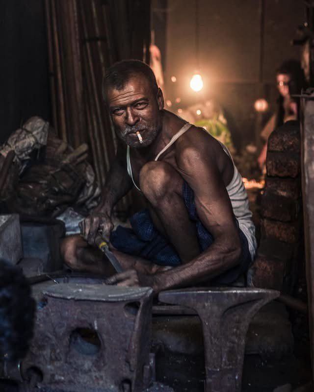 Portrait of a Blacksmith, Bangladesh: a man smoking, crouched in a dimly lit workshop. Photograph by Marcus Vallance.