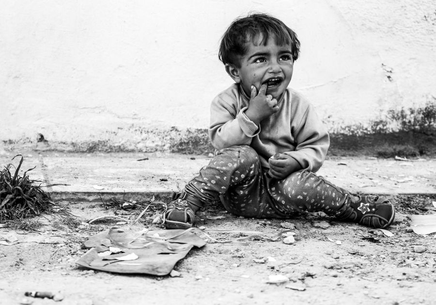 Portrait of a Girl, Greece: black and white photograph of a laughing toddler sitting on the ground by a white wall. Photograph by Marcus Vallance.