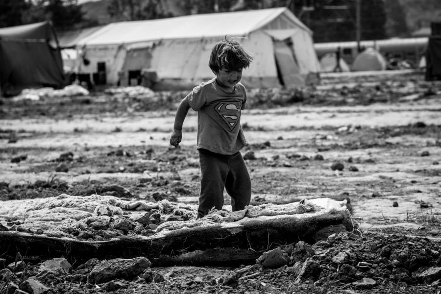Superman, Idomeni, Greece: black and white photograph of a small child in a superhero T-shirt walking across rough ground, tents behind. Photograph by Marcus Vallance.